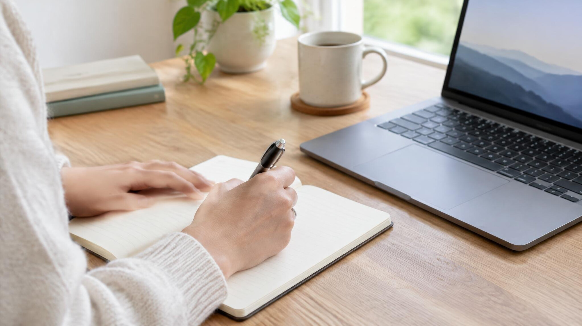 Person writing notes at a desk with a laptop and notebook